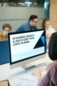 This is an image of a student on a Mac Desktop Computer. On the monitor, there is a bold graphic with a statement that says "Designers should always keep their users in mind." Below this statement in a smaller, light grey font, there is an additional statement that states, "Only then can you create truly innovative solutions."